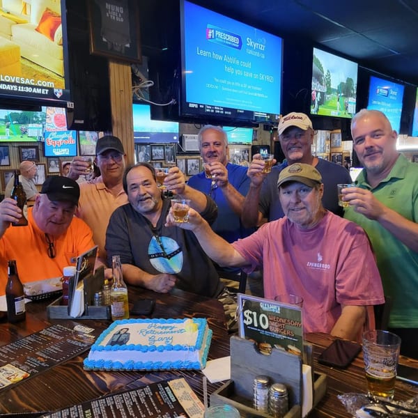 Seven men at Gleason's Bar raise their drinks, celebrating with a "Happy Retirement Gary" cake.