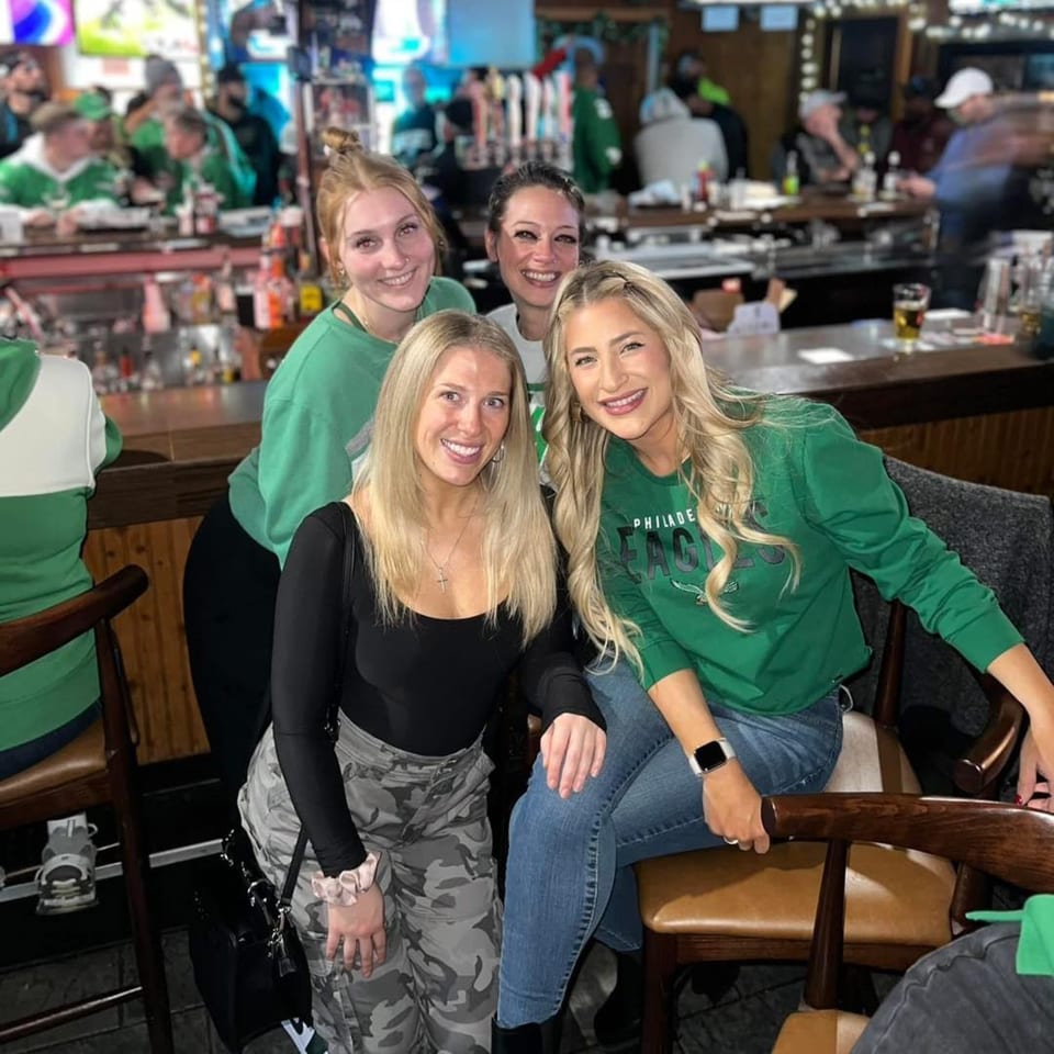 Four smiling women in green shirts and casual attire pose together inside a lively bar.
