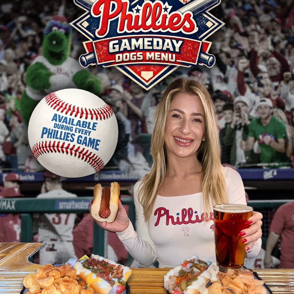 Woman in Phillies shirt holding a hot dog and beer, with two loaded hot dogs and chips.