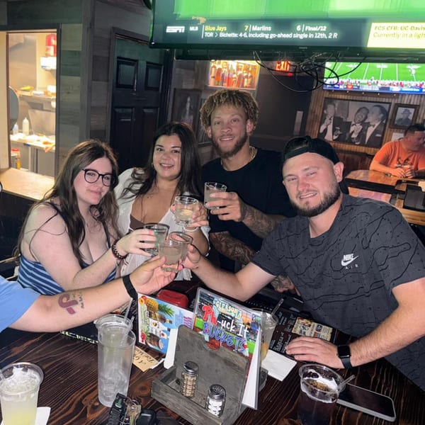 Four friends at Gleason's Bar clinking glasses, with a sports game on a TV in the background.