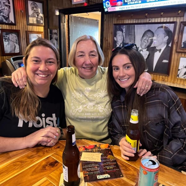 Three smiling women, two with drinks, pose at a wooden table inside Gleason's Bar.