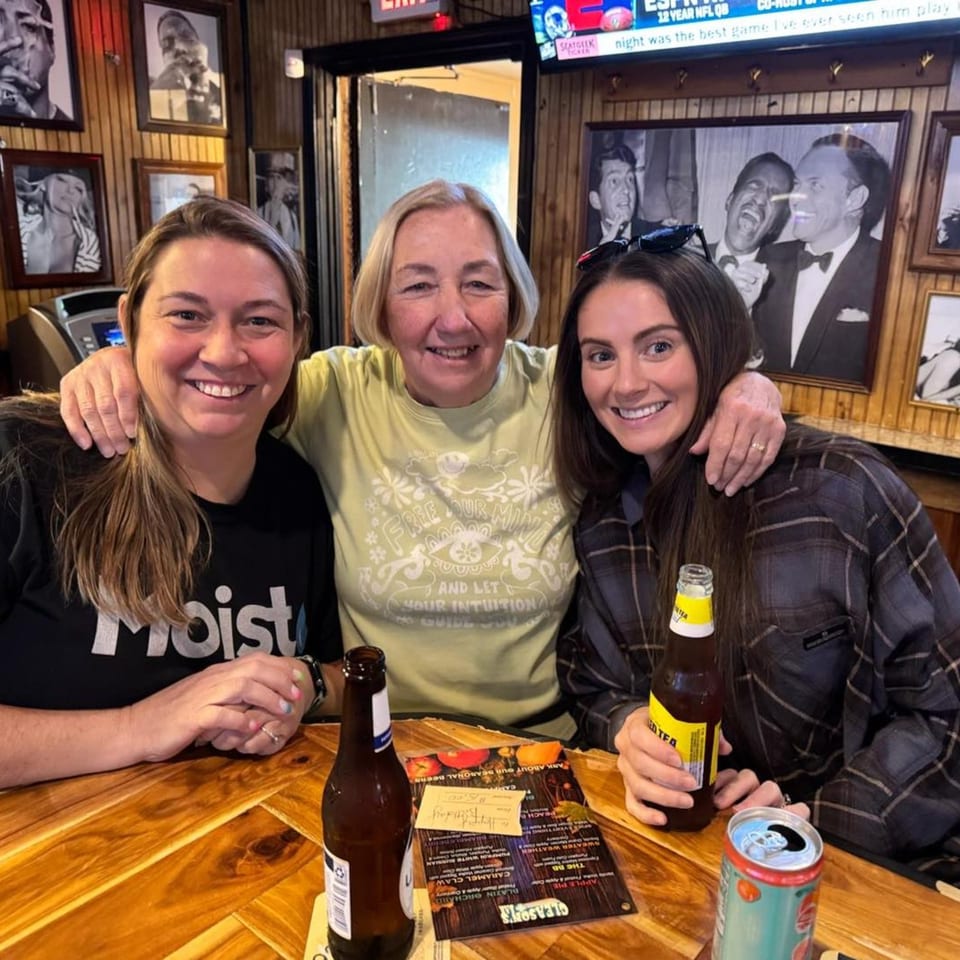 Three smiling women, two with drinks, pose at a wooden table inside Gleason's Bar.