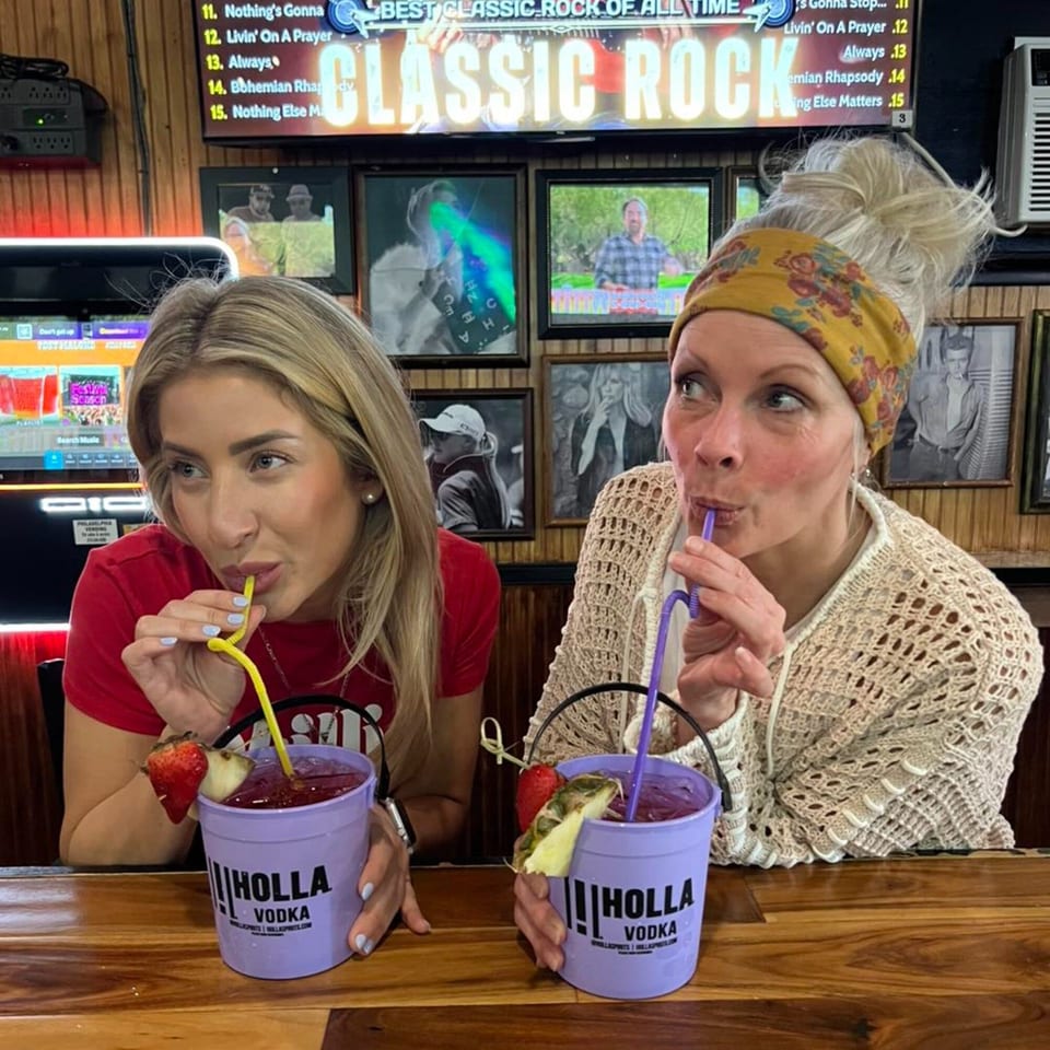 Two women at Gleasons Bar sip purple cocktails from "Holla Vodka" buckets with fruit garnishes.