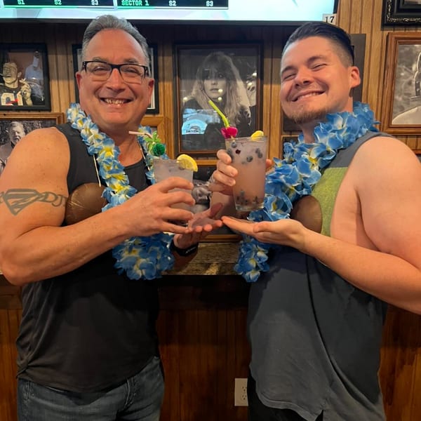 Two smiling men in Hawaiian leis and coconut bras hold blueberry cocktails at Gleasons Bar.